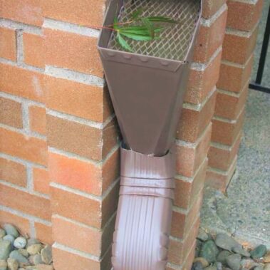 Brown gutter downspout with integrated mesh filter cleanout system installed on brick wall, showing effective leaf and debris collection