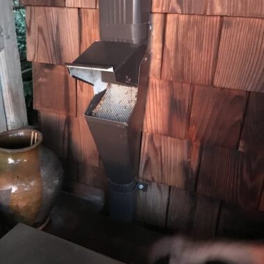 Gutter cleanout with protective mesh cover installed on cedar siding, preventing debris buildup while maintaining easy maintenance access