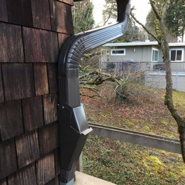Brown gutter system with protective cleanout cover and curved downspout installed on cedar siding, featuring leaf protection for Pacific Northwest homes