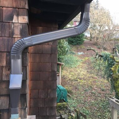 Brown curved gutter system with special cleanout cover installed on wooden shingle house, showcasing efficient water flow and easy maintenance design