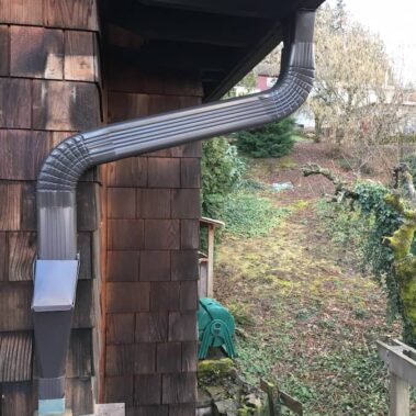 Brown corrugated downspout with easy-access cleanout lid installed on cedar shake siding, featuring practical maintenance design by Royal Gutters Portland