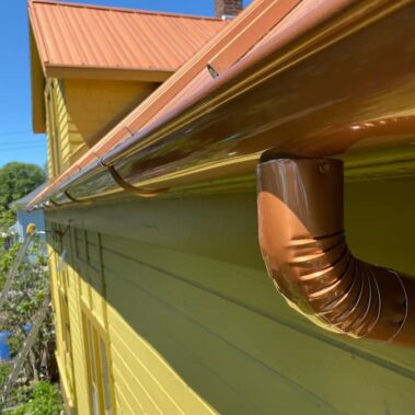 Close-up of copper metallic half-round gutter with round downspout, showing brilliant copper finish and expert craftsmanship against yellow siding