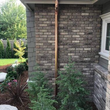 Bright copper round downspout installed on brick wall, showing clean professional installation and natural copper finish against grey brick exterior