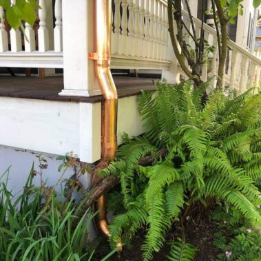 Gleaming copper round downspout on white Victorian porch, complementing ornate railings and lush fern landscaping, showcasing timeless elegance