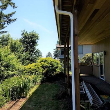 White corrugated downspout professionally installed on modern home, showing clean vertical installation with proper landscaping drainage integration